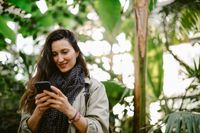 Frau im Freien betrachtet lächelnd ein Mobiltelefon, umgeben von Bäumen.